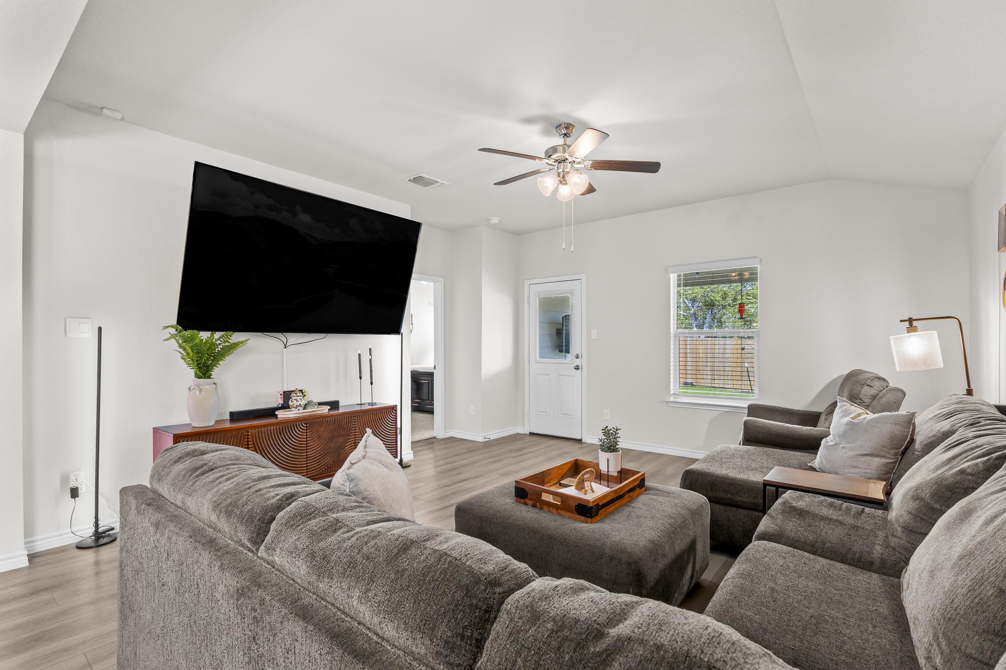 Furnished living room with a gray sectional, large wall-mounted television, and ceiling fan.