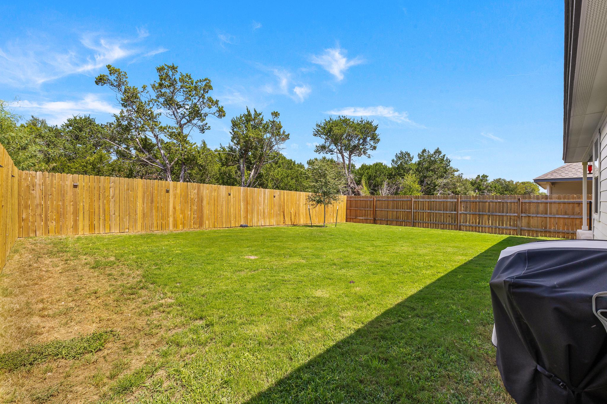 Fenced backyard with a broad green lawn, mature trees, and a covered grill.