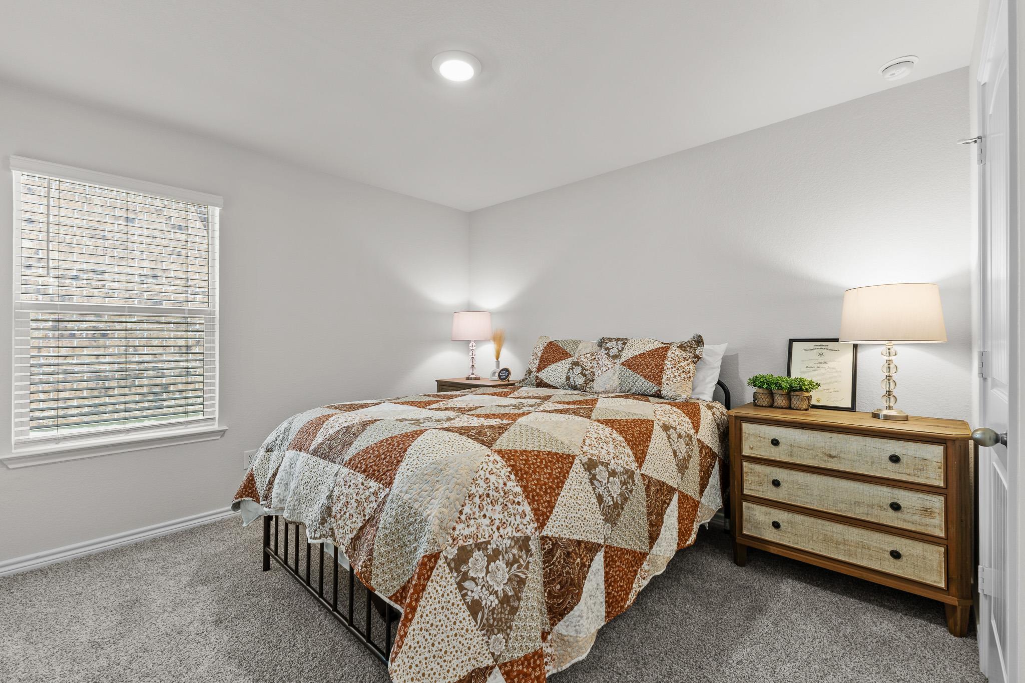 Furnished bedroom with a patchwork quilt, wooden dresser, and window with blinds.