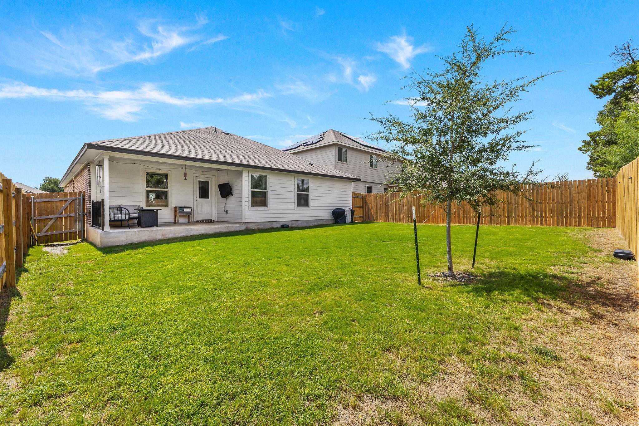 House rear and covered patio overlooking a fenced backyard with a young tree.