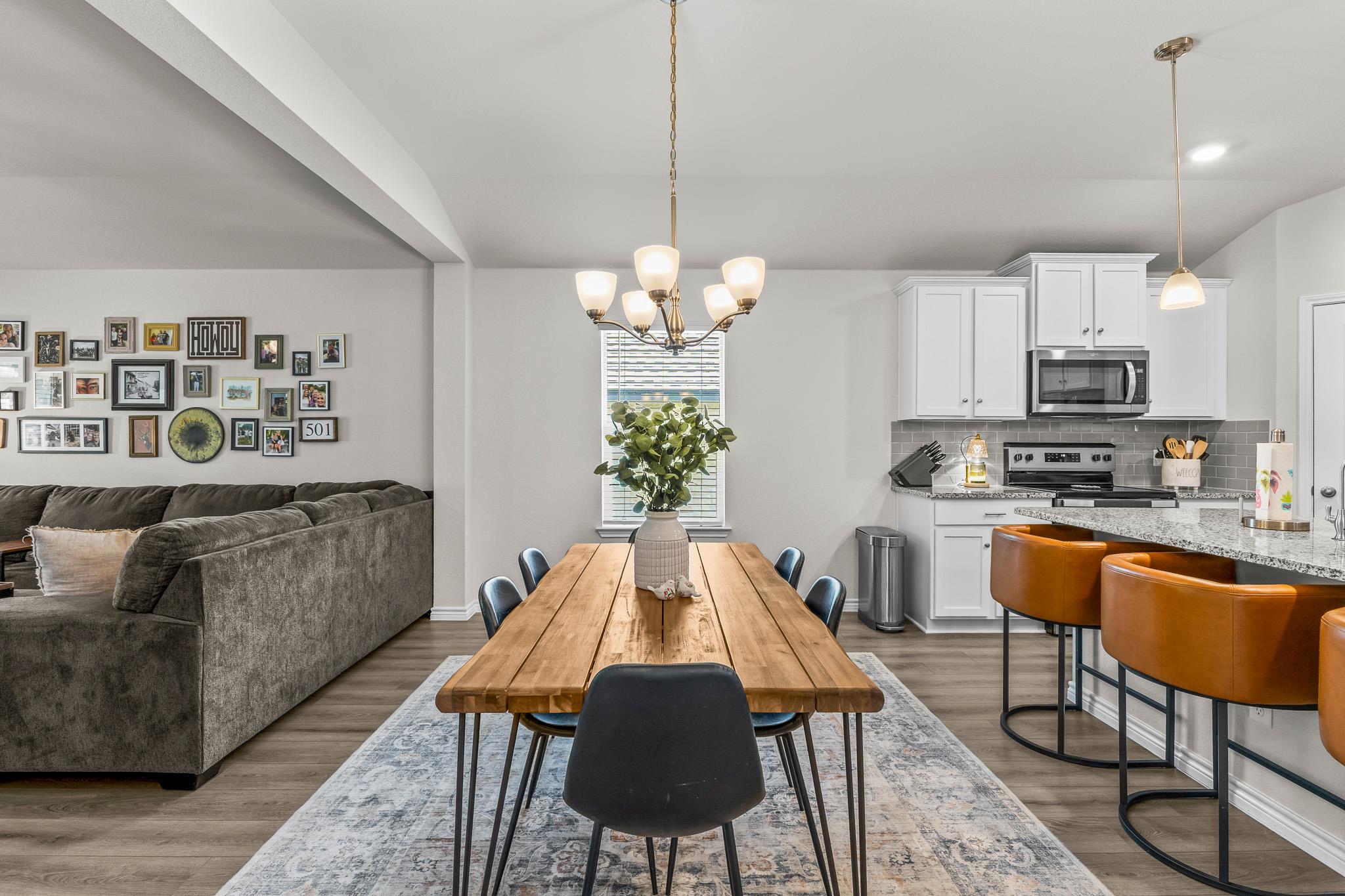 Open-plan dining area with a long wooden table, gray sectional, and white kitchen with breakfast bar.