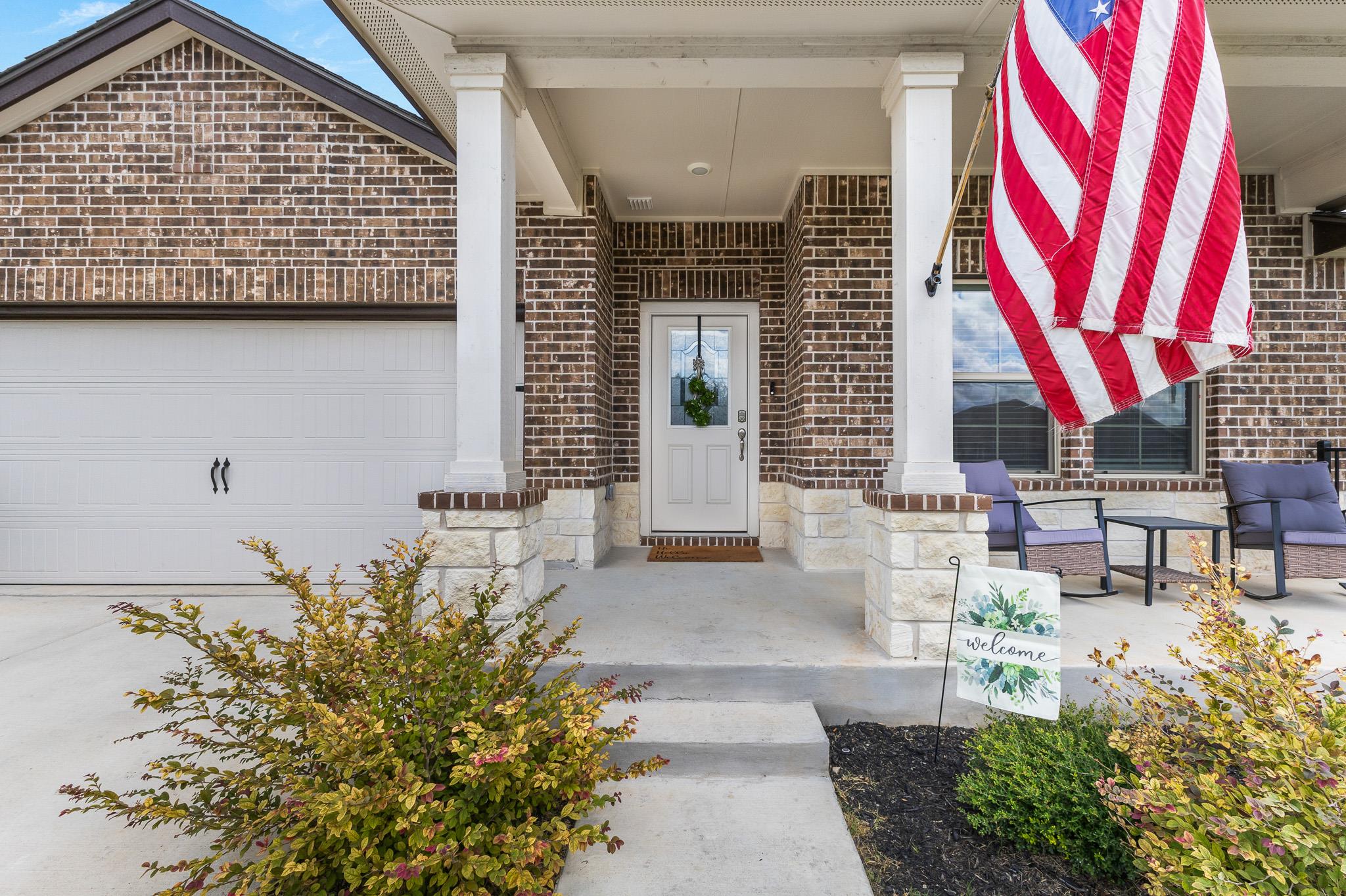 Brick home entrance with a covered porch, American flag, and landscaped shrubs.