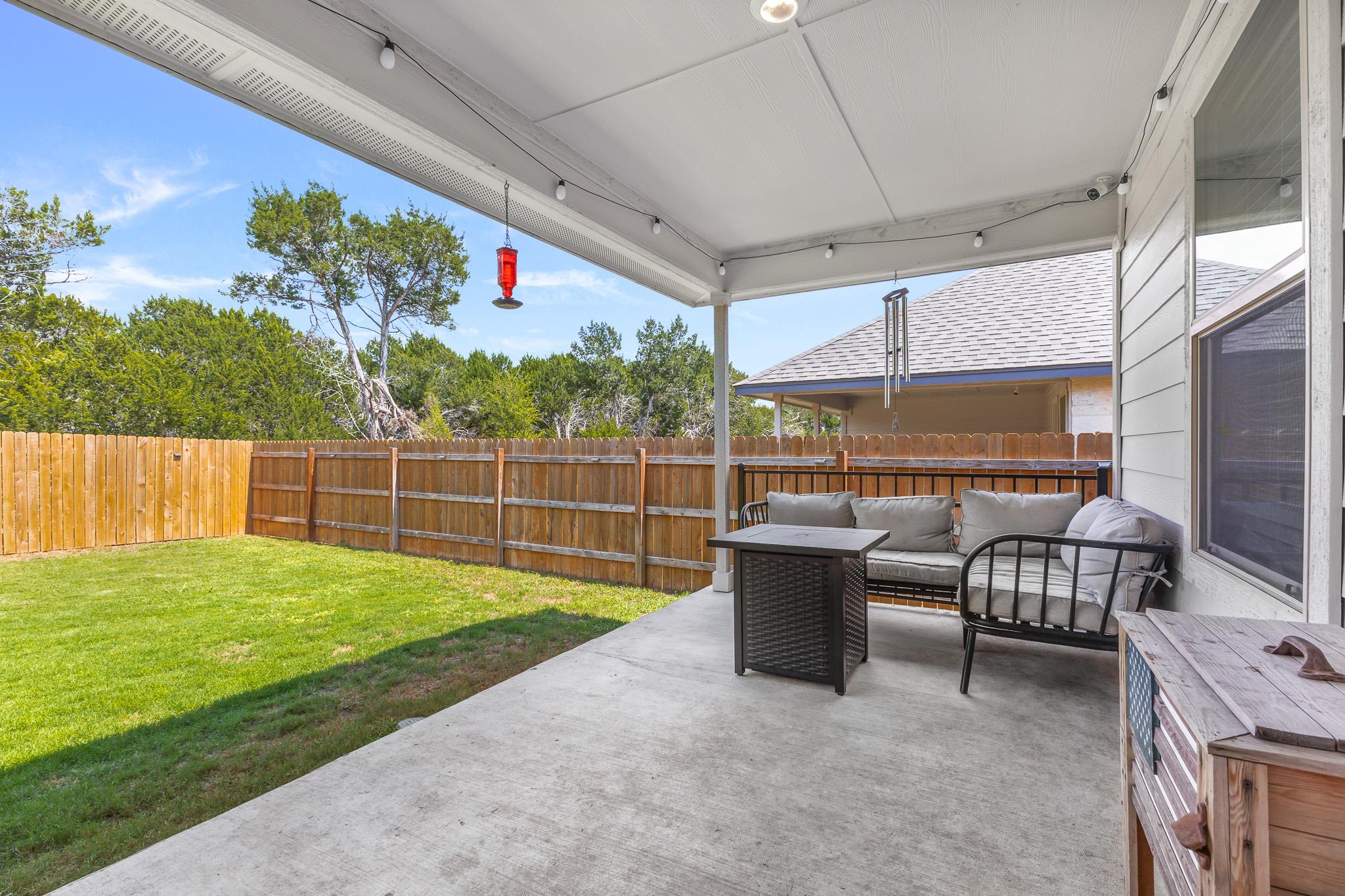 Covered patio with cushioned seating and a wooden storage chest beside a fenced grassy backyard.