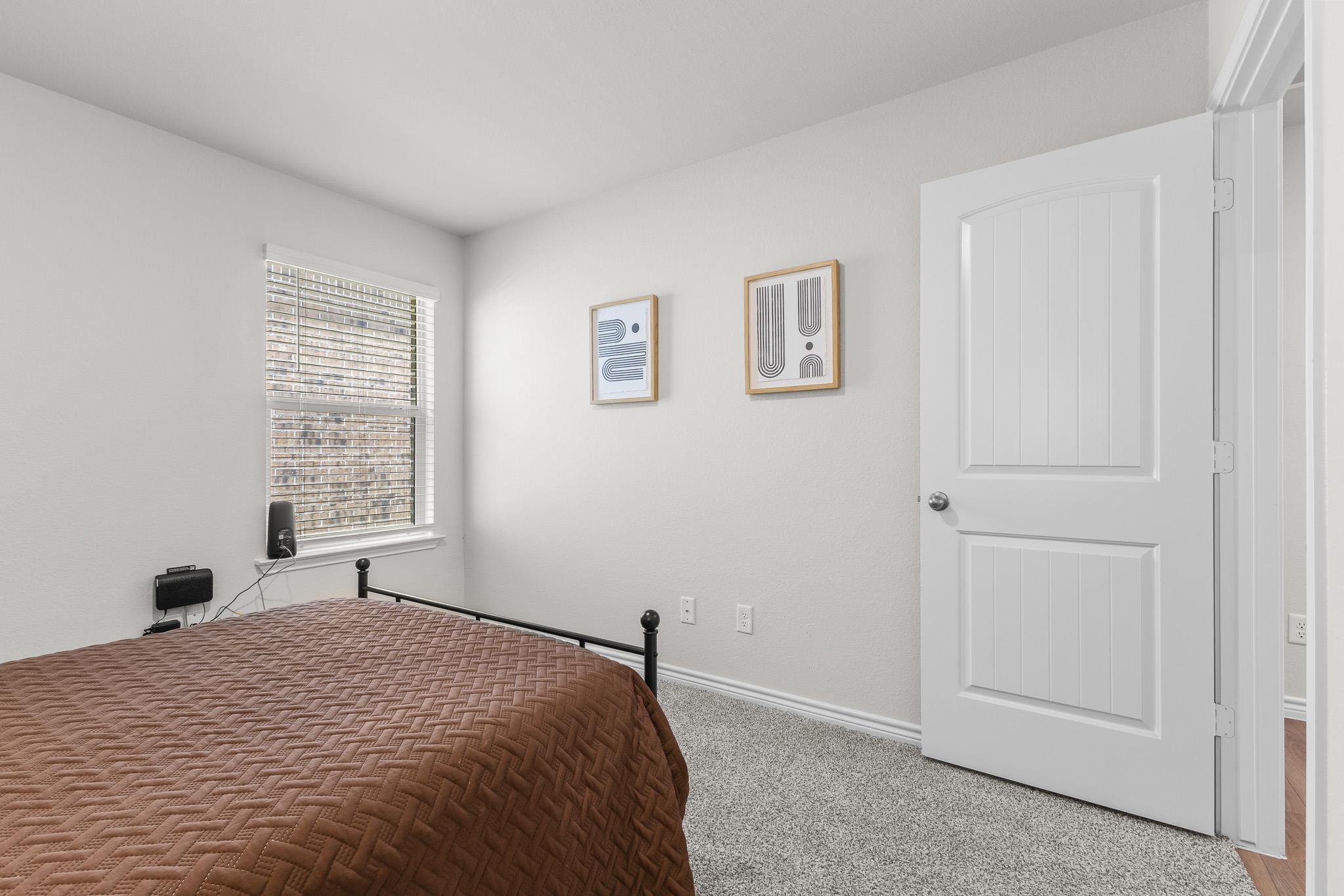 Bedroom with a brown quilted bed, window blinds, framed prints, and an open white door.