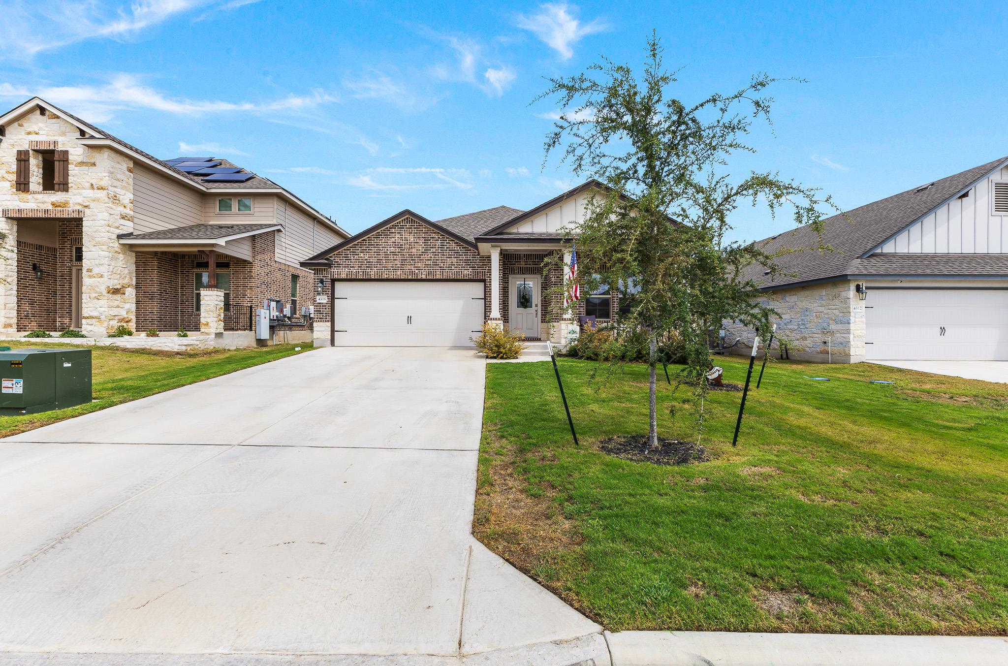 Brick house with a two-car garage, wide driveway, and young tree in the front yard.