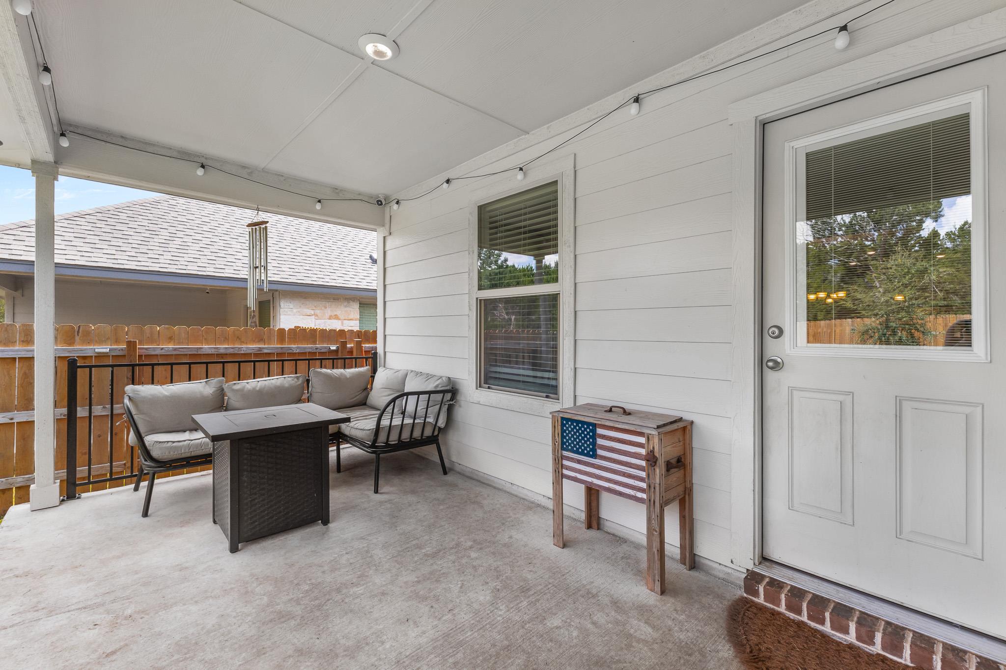 Covered back patio with cushioned seating, string lights, and a wooden fence beyond the railing.