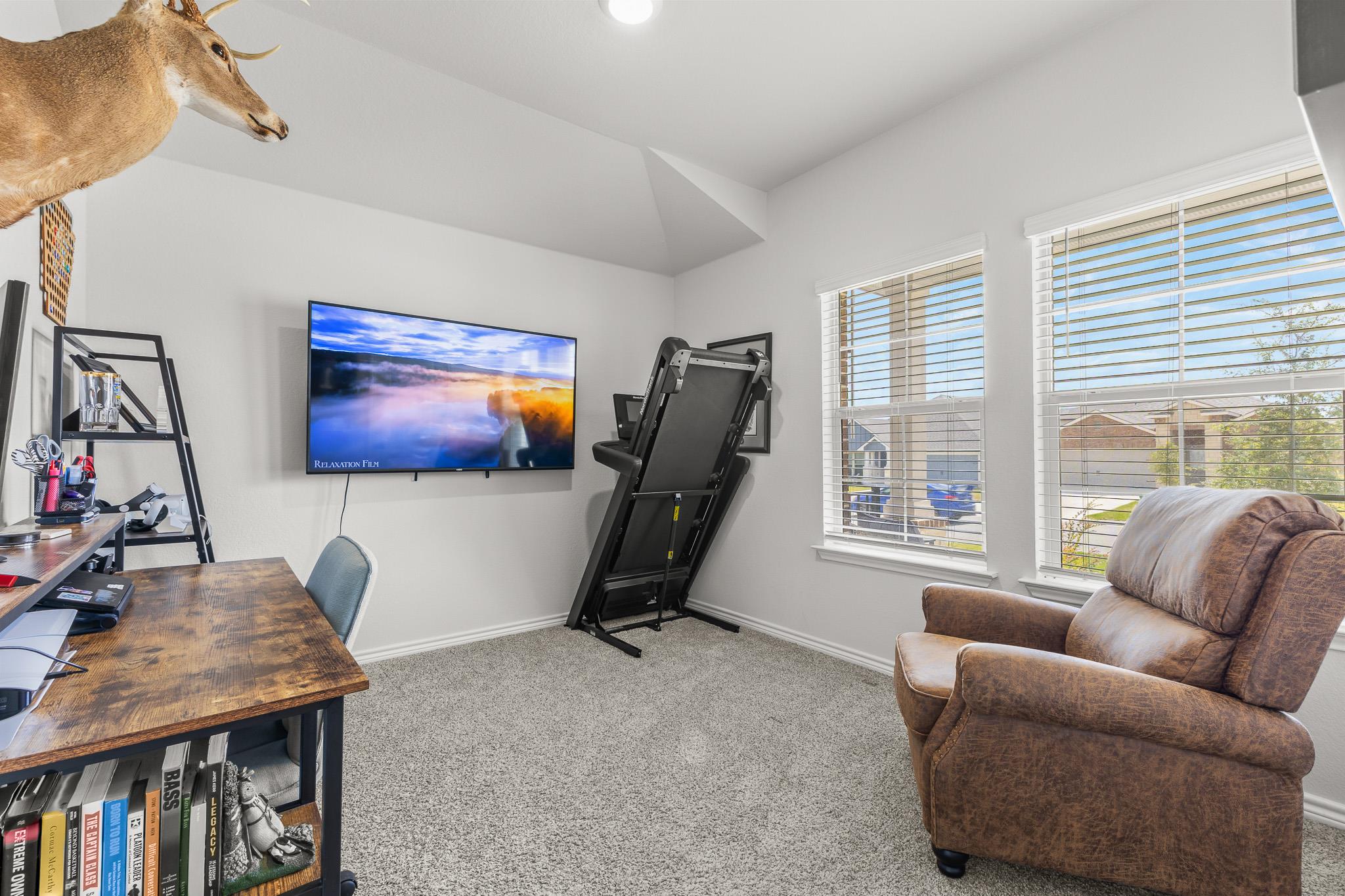Home office and exercise room with a treadmill, wall-mounted TV, wooden desk, and brown recliner.