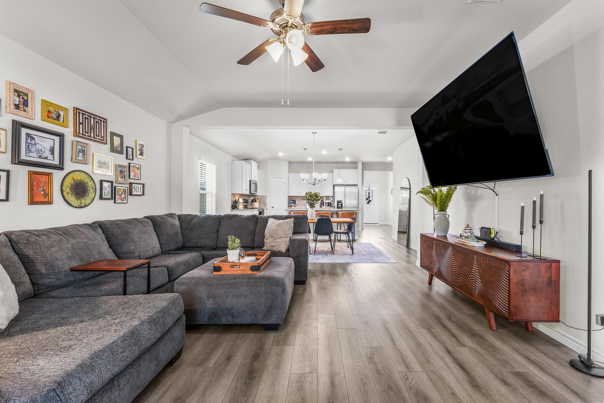Open-plan living room with a gray sectional, gallery wall, and kitchen and dining area beyond.