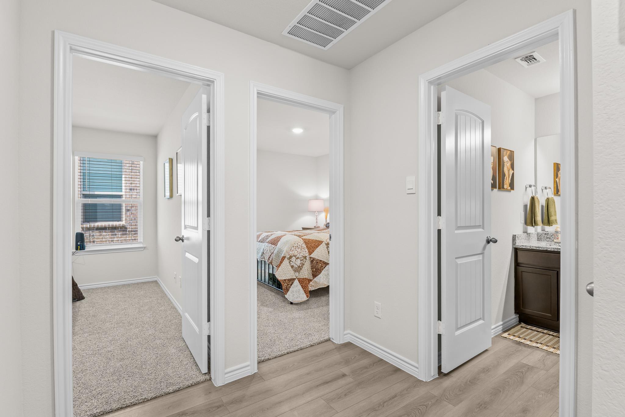 Hallway with open doors to a bedroom, carpeted room, and bathroom with dark vanity.