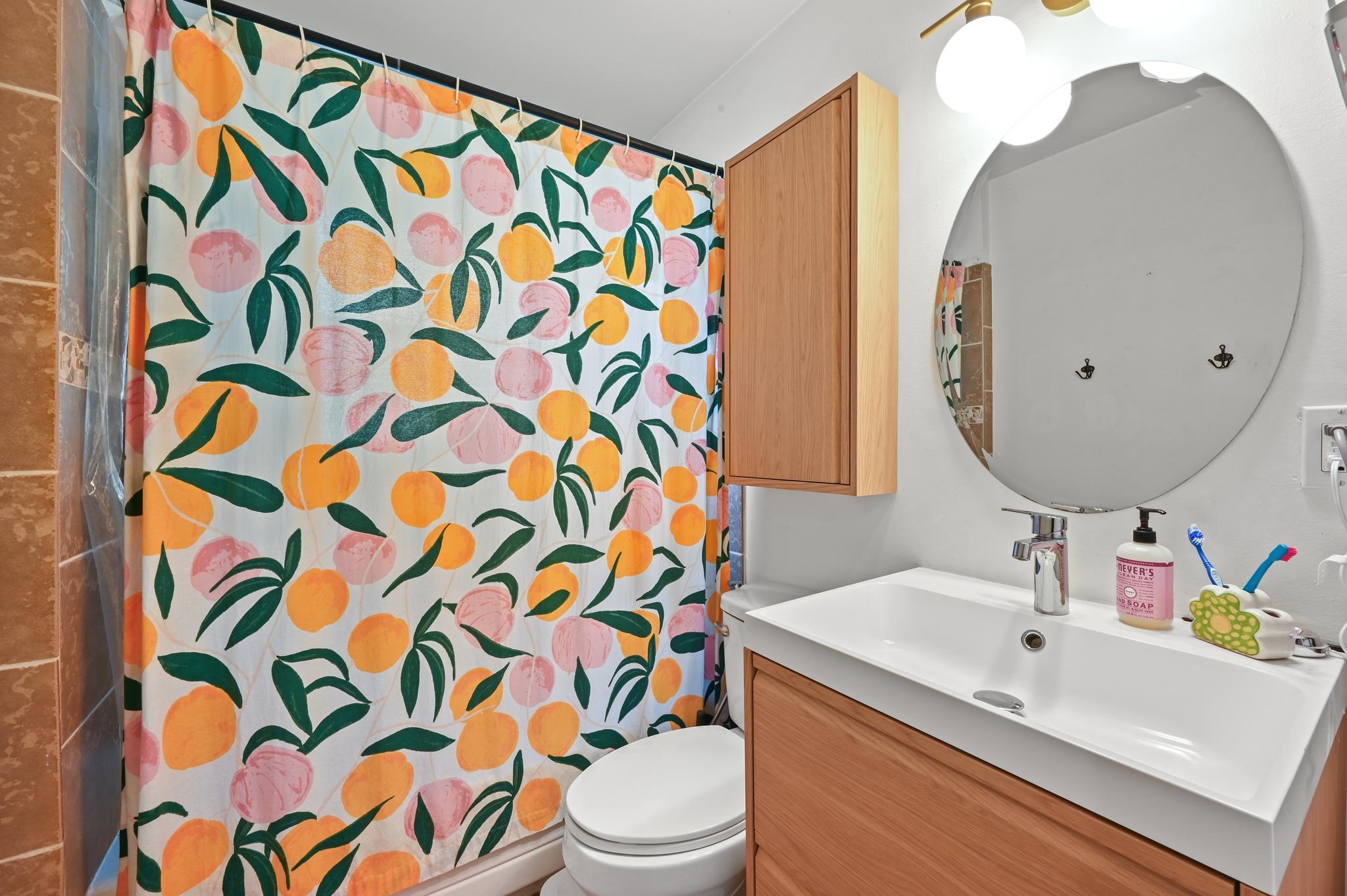 Bathroom with a fruit-print shower curtain, white sink, round mirror, and warm wood cabinetry.