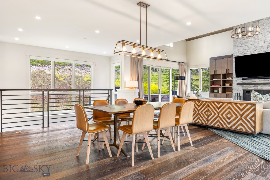 Open-plan dining and living area with a wood dining table, large windows, and stone fireplace.