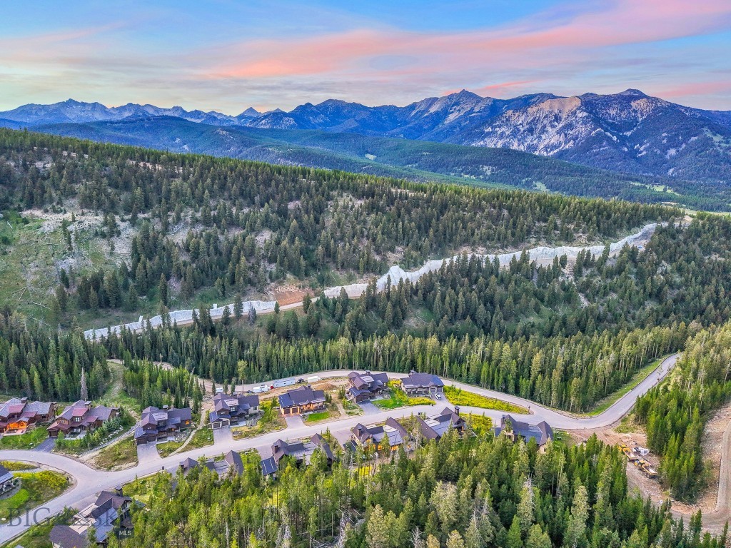 Aerial view of a mountain neighborhood surrounded by forest, with a distant range beneath a pink evening sky.