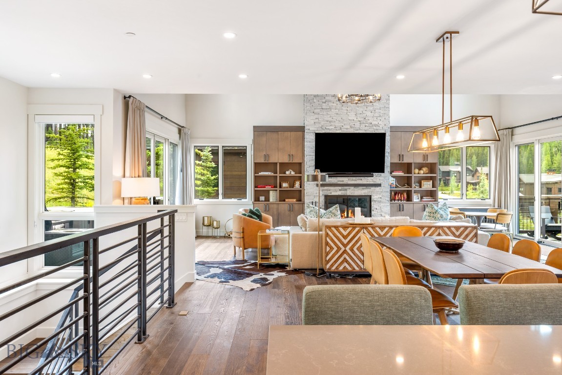 Open-plan living and dining room with a stone fireplace, built-ins, and expansive windows overlooking trees.