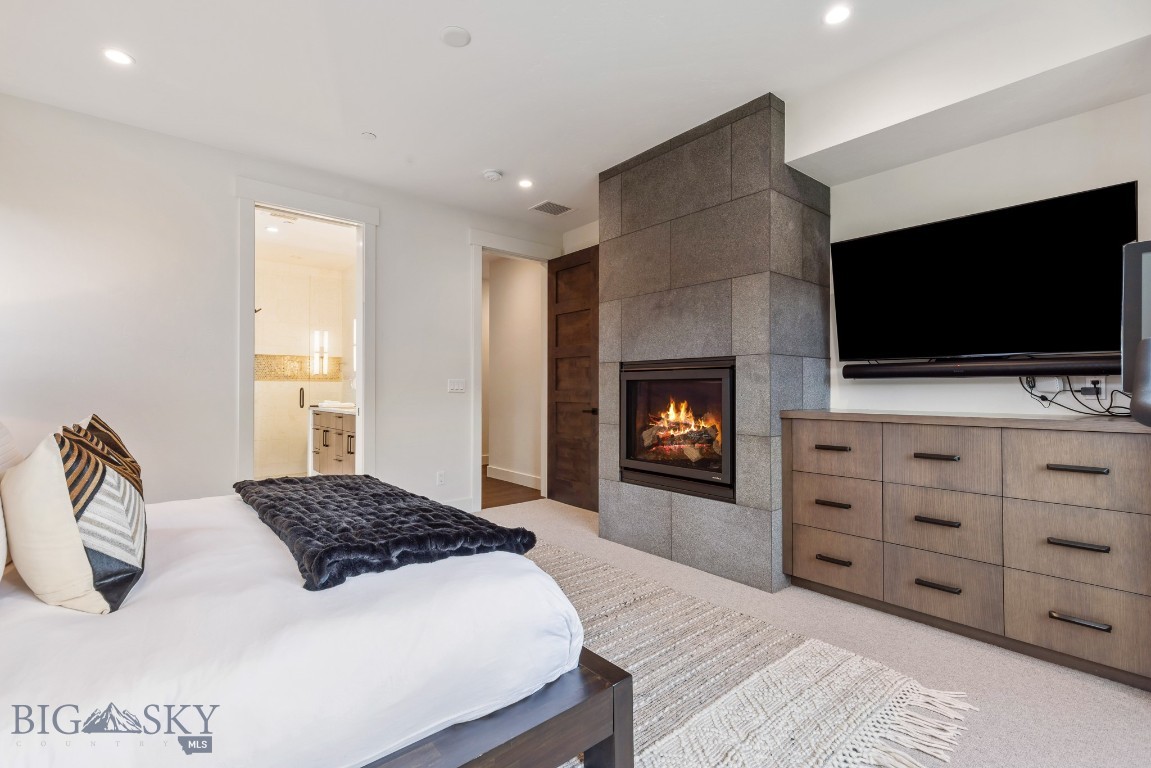 Bedroom with a stone fireplace, lit fire, wall-mounted television, and white bed.