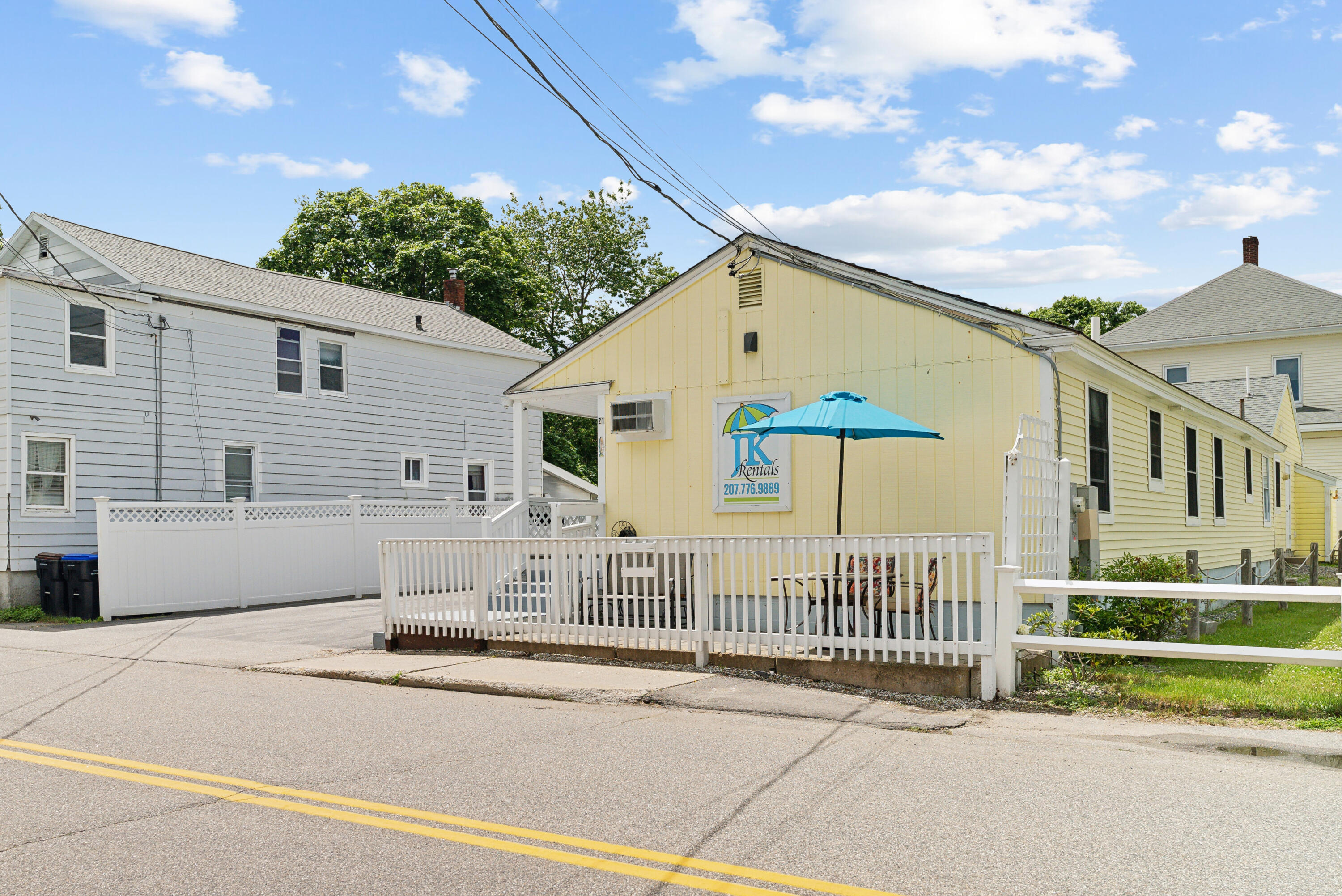 Yellow cottage with a white-railed deck and turquoise patio umbrella beside the street.