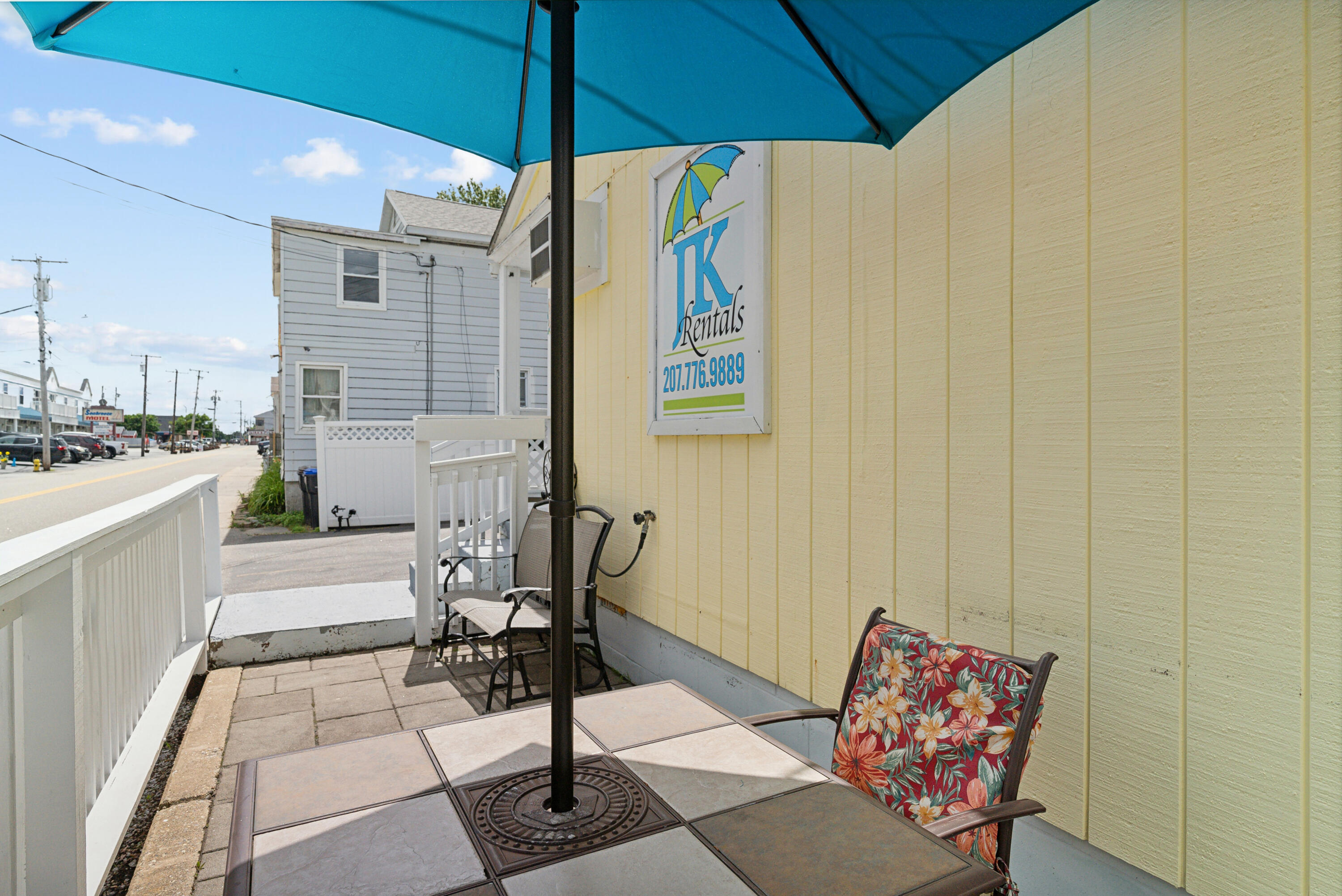 Patio with a blue umbrella, outdoor seating, and an LK Rentals sign beside the street.
