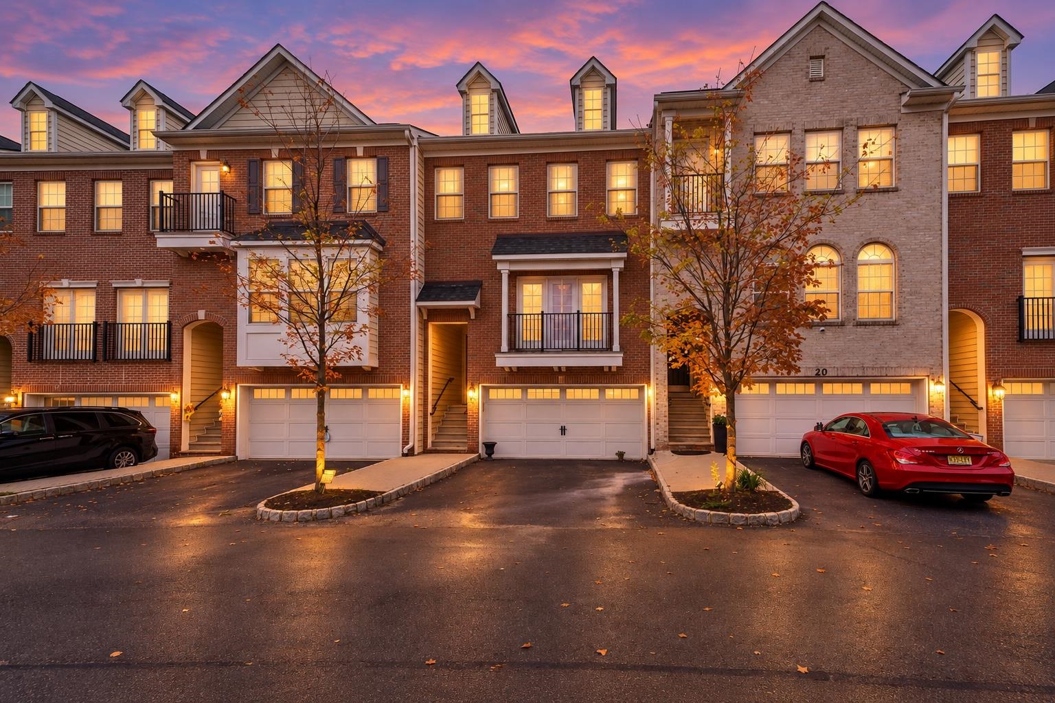 Attached brick townhouses at dusk, with illuminated windows, autumn trees, and cars parked by the garages.