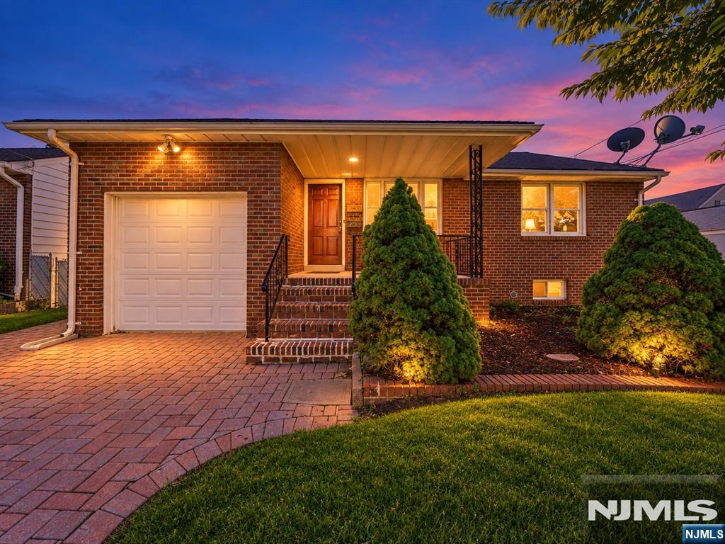 Brick house at dusk with a white garage door, paver driveway, and landscaped front yard.