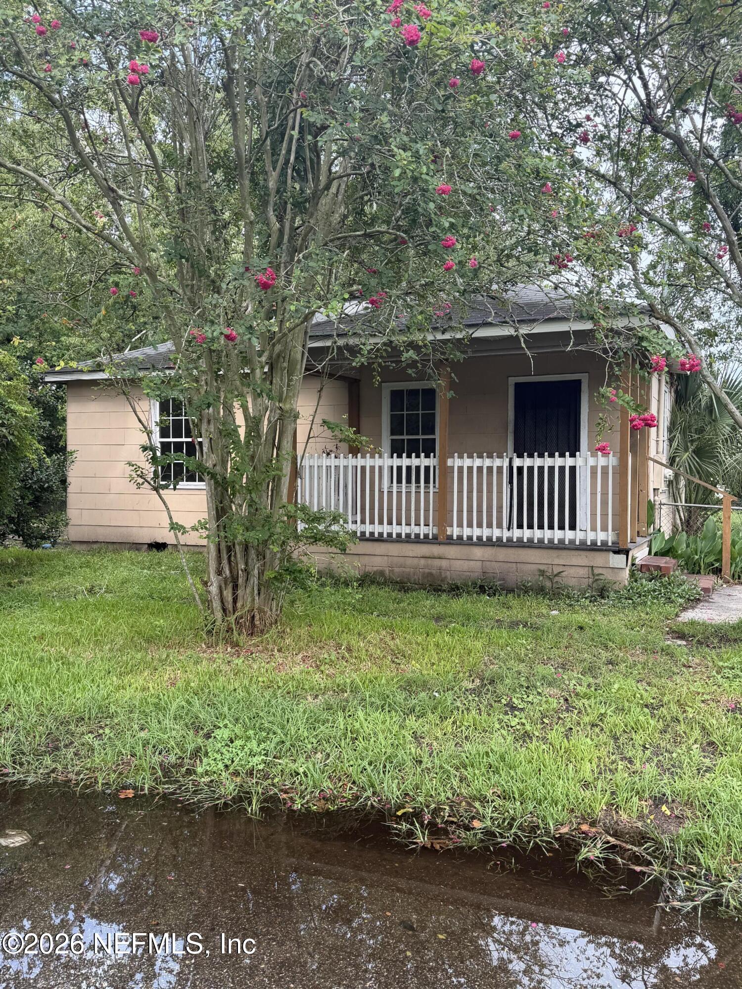 Small tan house with a white-railed porch behind a flowering tree, with a creek in the foreground.