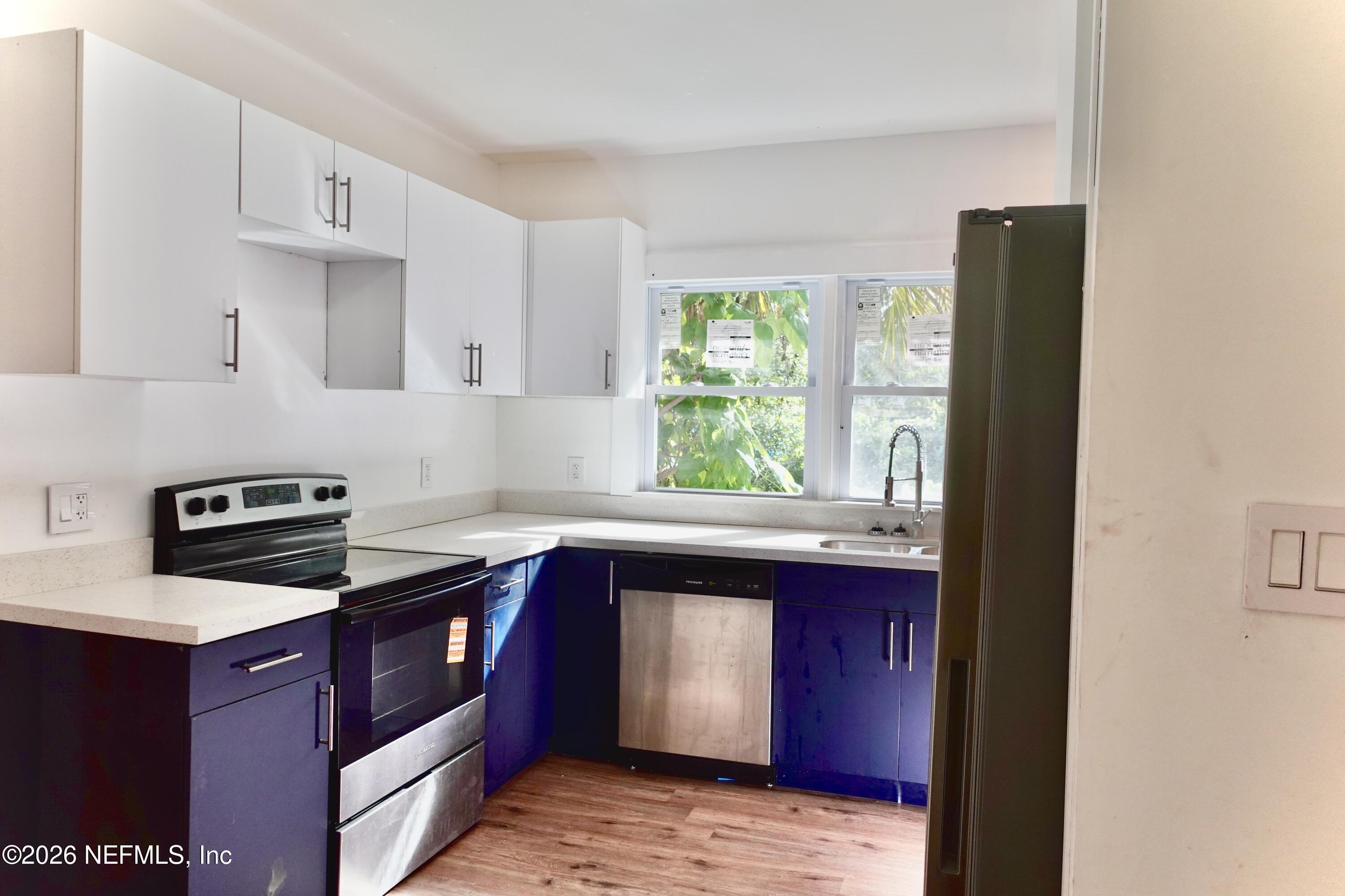Kitchen with blue lower cabinets, stainless steel appliances, and a sink beneath two windows.