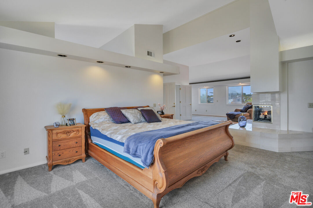 Staged bedroom with carved wooden bed, gray carpet, and fireplace opening to a sitting area.