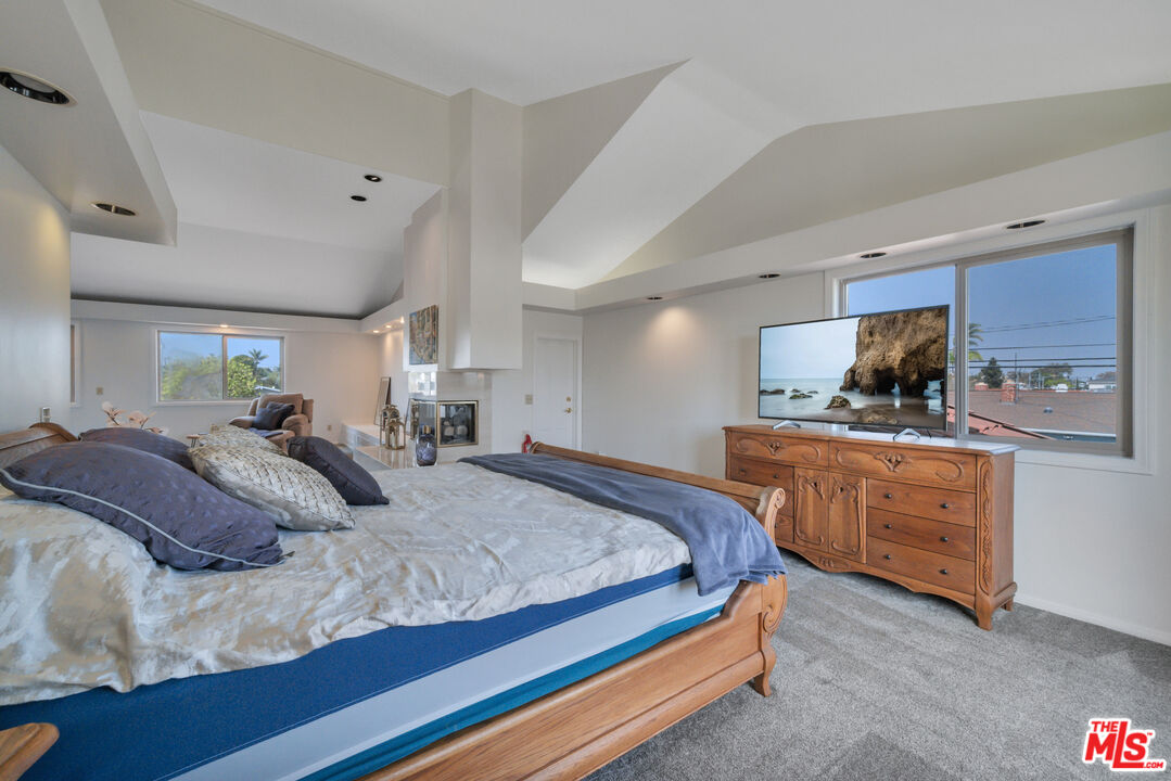 Furnished bedroom with a large wooden bed, fireplace, and television beside broad windows.