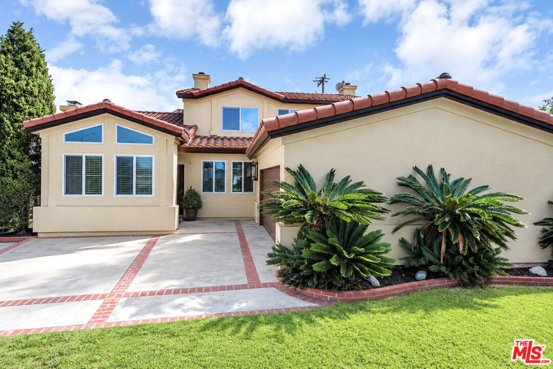 Beige two-story home with a red tile roof, brick-edged driveway, cycads, and front lawn.
