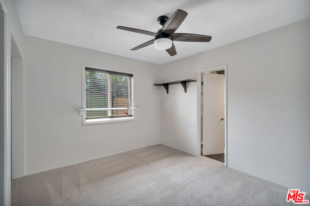 Empty carpeted bedroom with a window, ceiling fan, and small wall shelf.