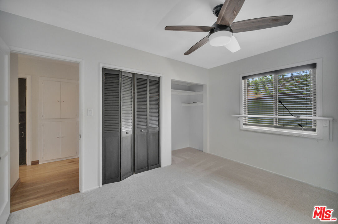 Vacant bedroom with dark louvered closet doors, a ceiling fan, and a window overlooking greenery.