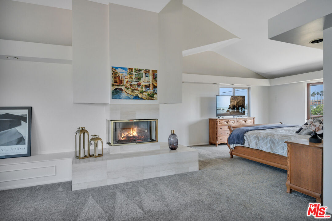 Bedroom with a vaulted ceiling, lit marble fireplace, and wooden bed beside tall windows.
