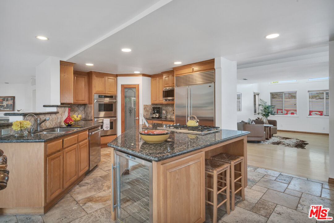 Kitchen with a granite-topped island, warm wood cabinetry, and stainless steel appliances opening to a living area.
