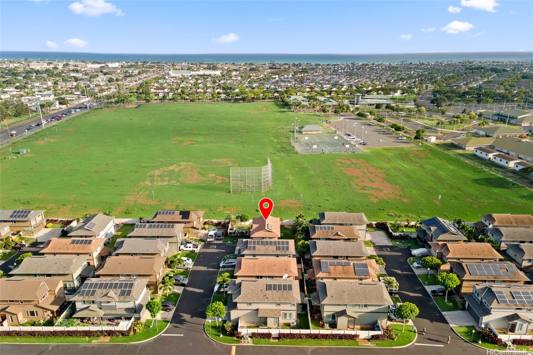 Aerial view of the marked Ewa Beach home beside a large green field, with tennis courts and ocean beyond.