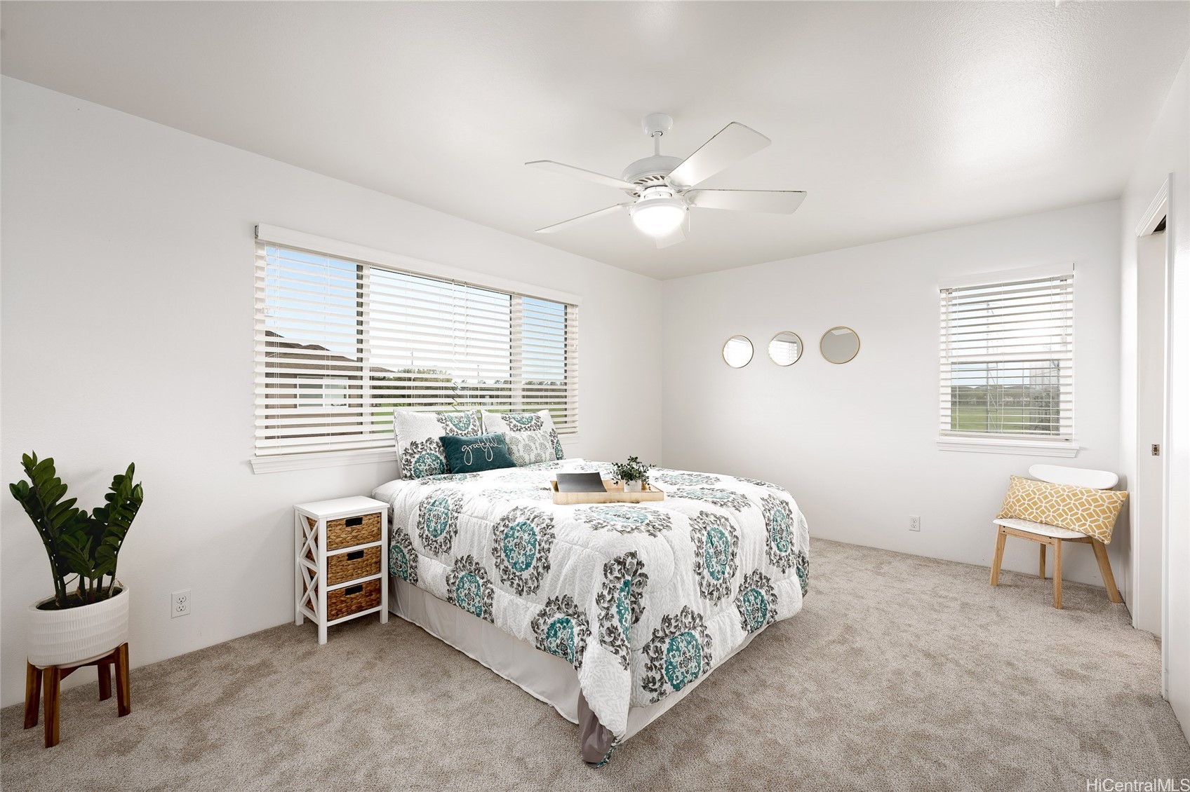 Staged bedroom with patterned bedding, a ceiling fan, round mirrors, and a chair beside the window.