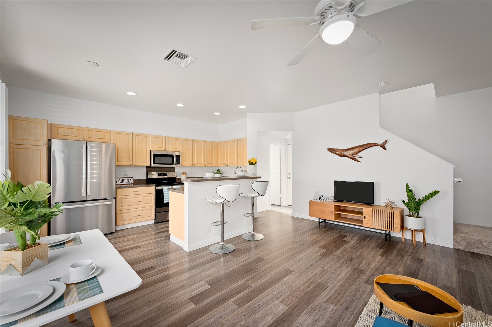 Open-plan kitchen and living area with light wood cabinetry, stainless appliances, breakfast bar, and whale wall decoration.