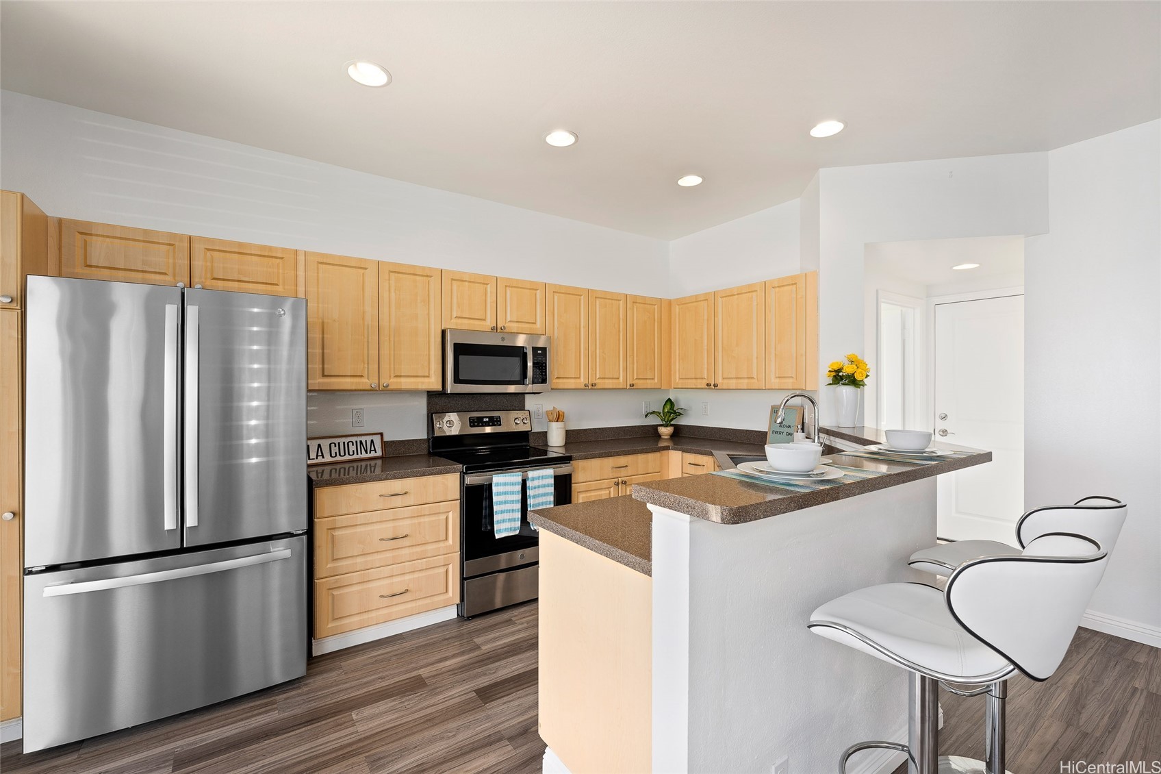 Kitchen with light wood cabinets, stainless steel appliances, dark counters, and a peninsula with white bar stools.