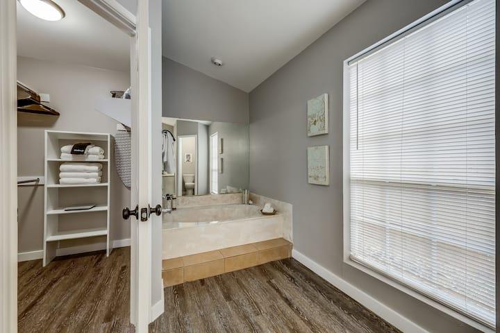 Bathroom with a raised tiled soaking tub, large mirrored vanity area, and window with white blinds.