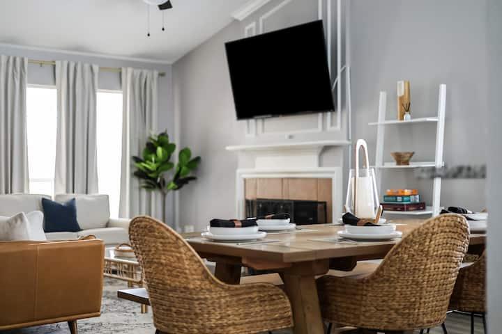 Living and dining room with woven chairs, a fireplace, and tall curtained windows.