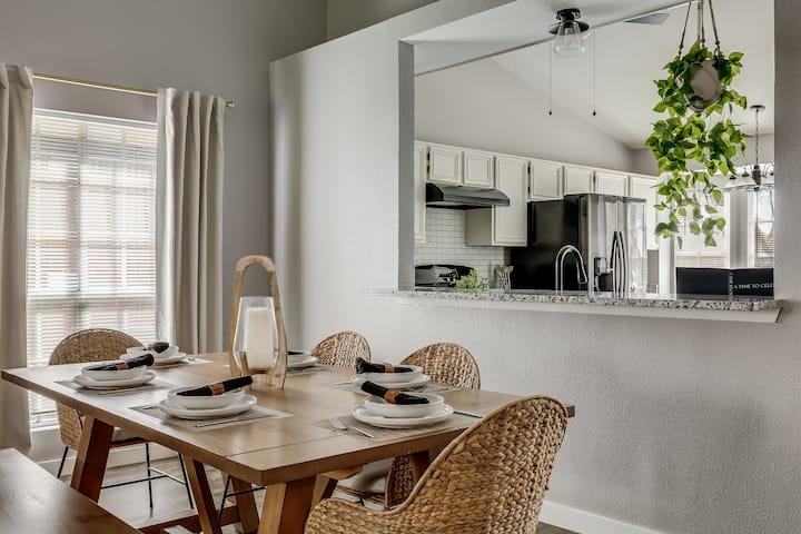 Dining area with a set wooden table and woven chairs overlooking a bright white kitchen.