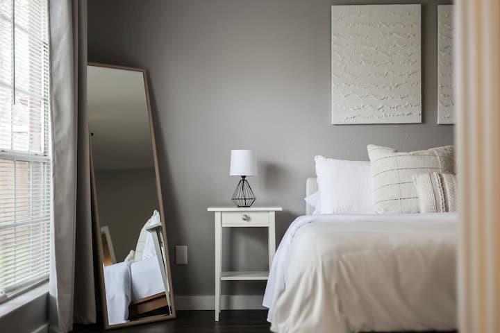 Staged bedroom with a white bed, bedside lamp, leaning mirror, and window with blinds.