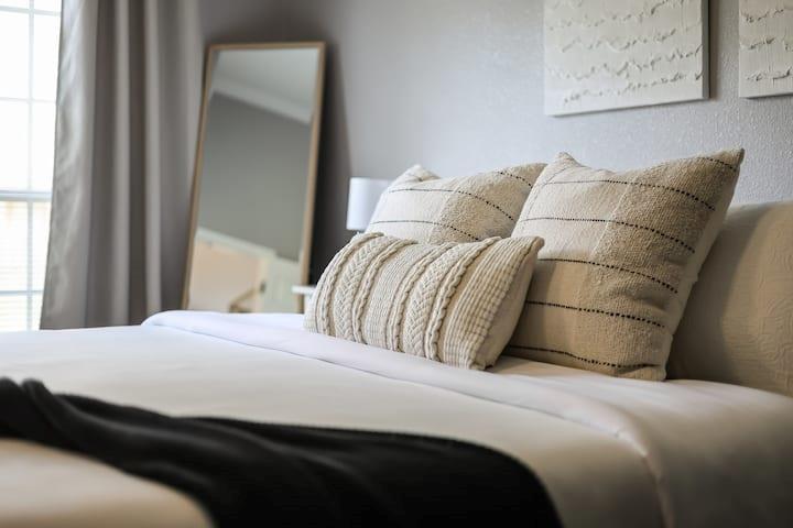 Staged bedroom with a white bed, textured cream pillows, black throw, and leaning mirror.