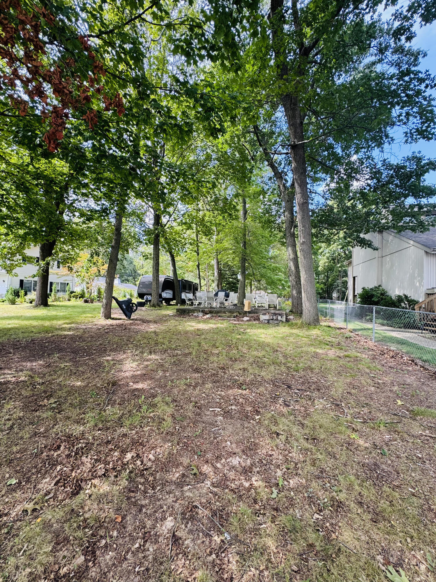 Shaded yard with mature trees, outdoor seating, and a travel trailer beneath the canopy.