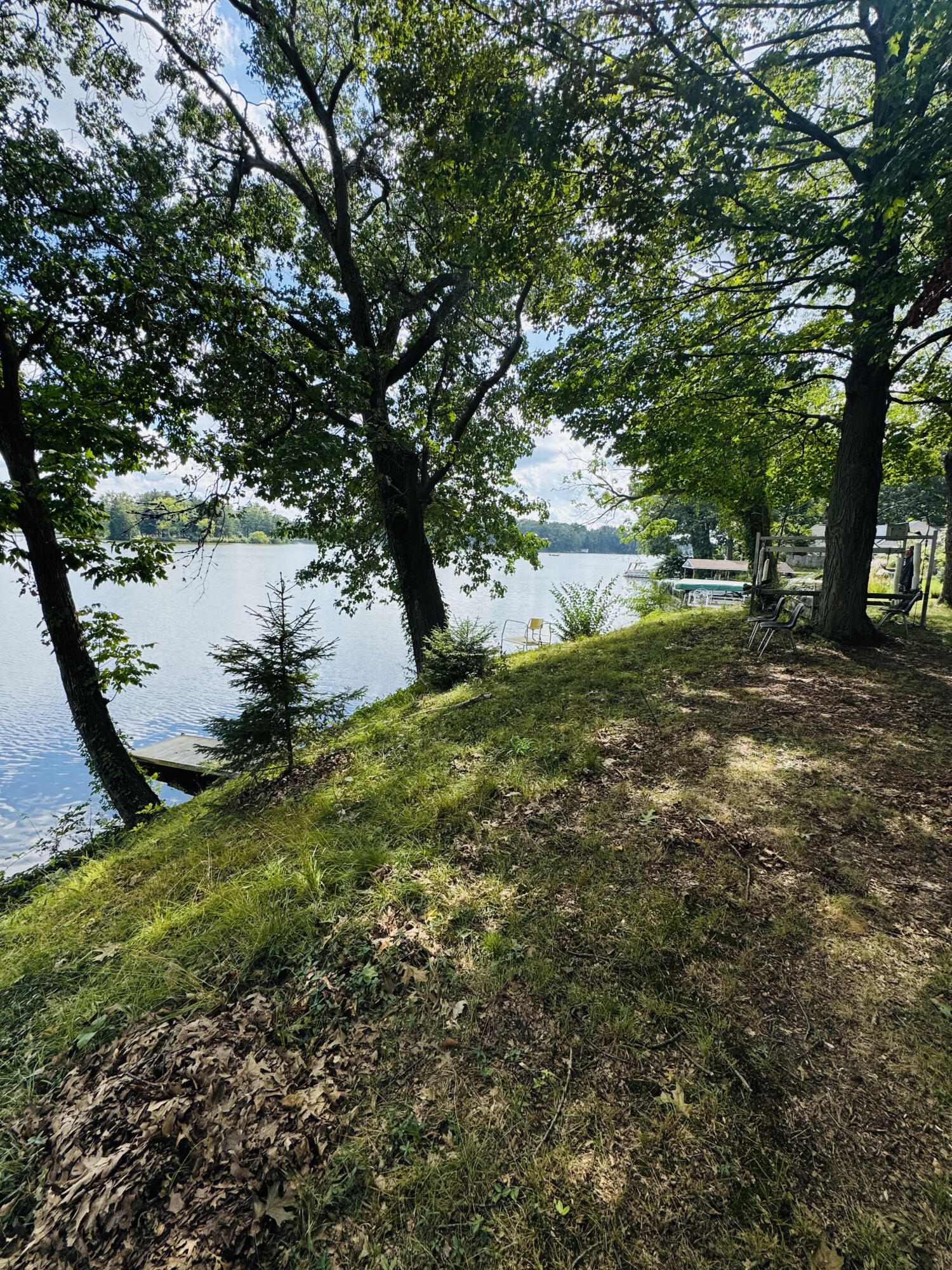 Shaded grassy lakeshore with a small dock, chairs, and boats on the water.