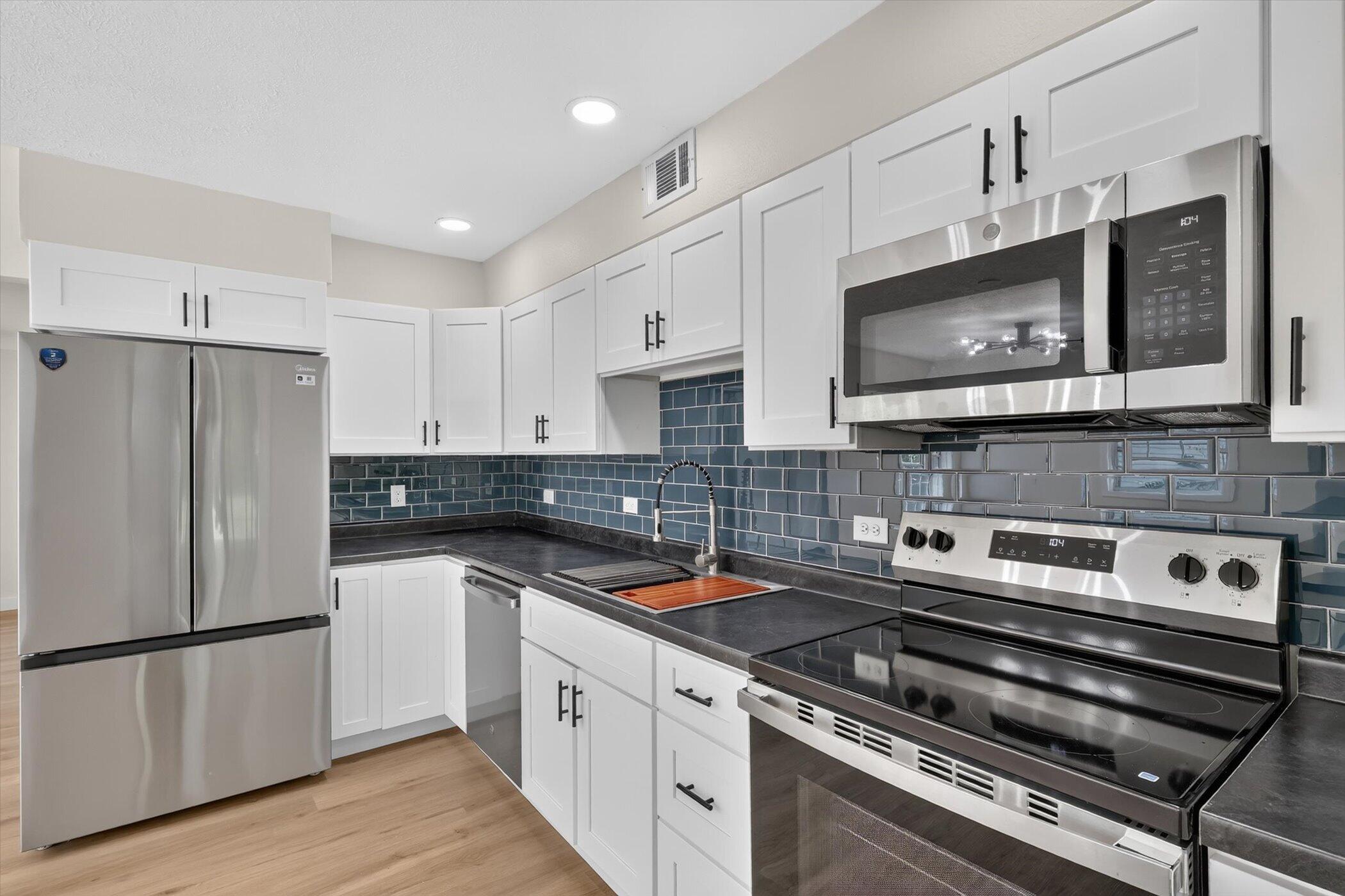 Kitchen with white shaker cabinets, blue-gray tile backsplash, dark countertops, and stainless steel appliances.