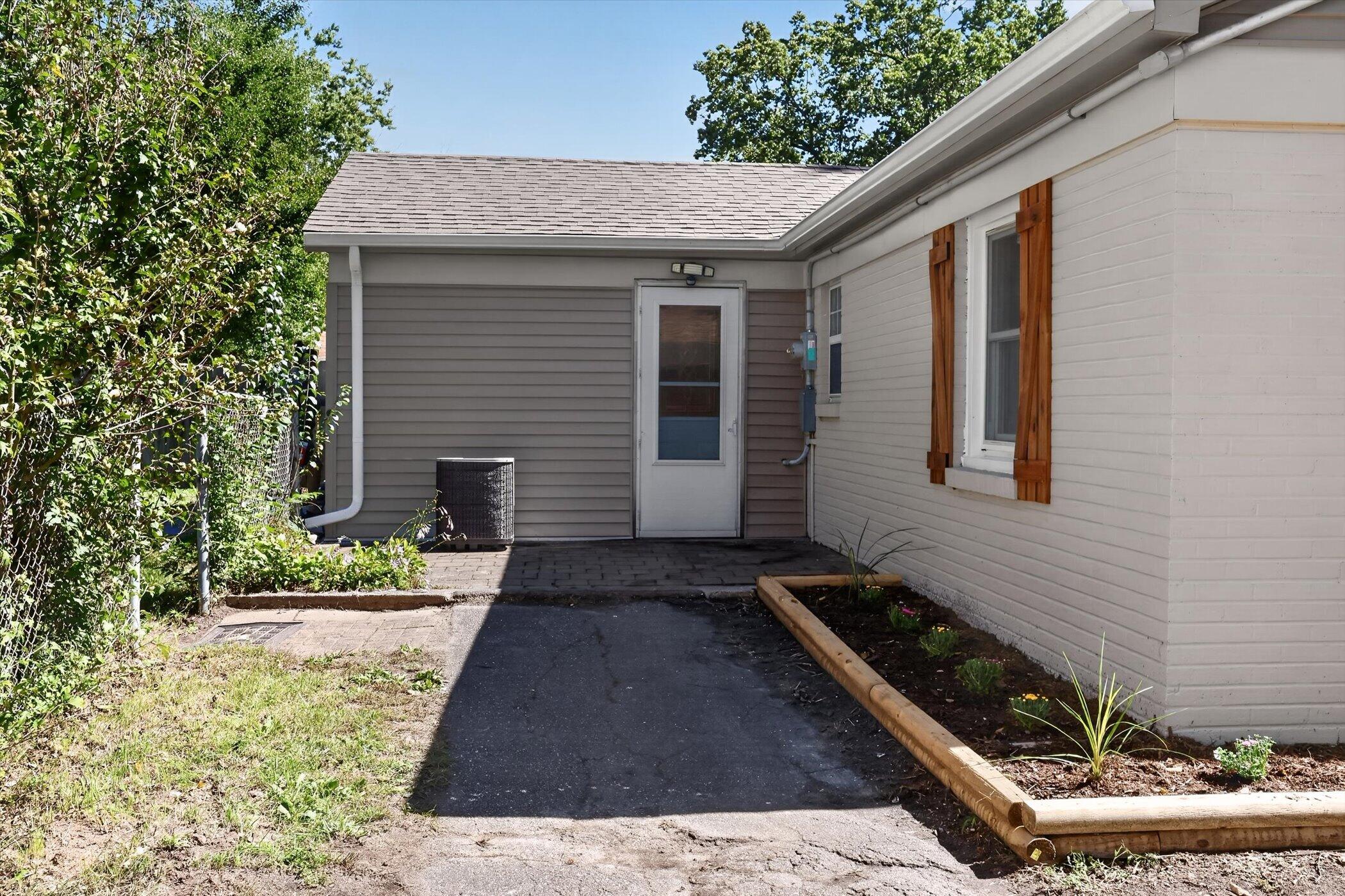 House side entrance with gray siding, wood window shutters, driveway, and raised garden bed.