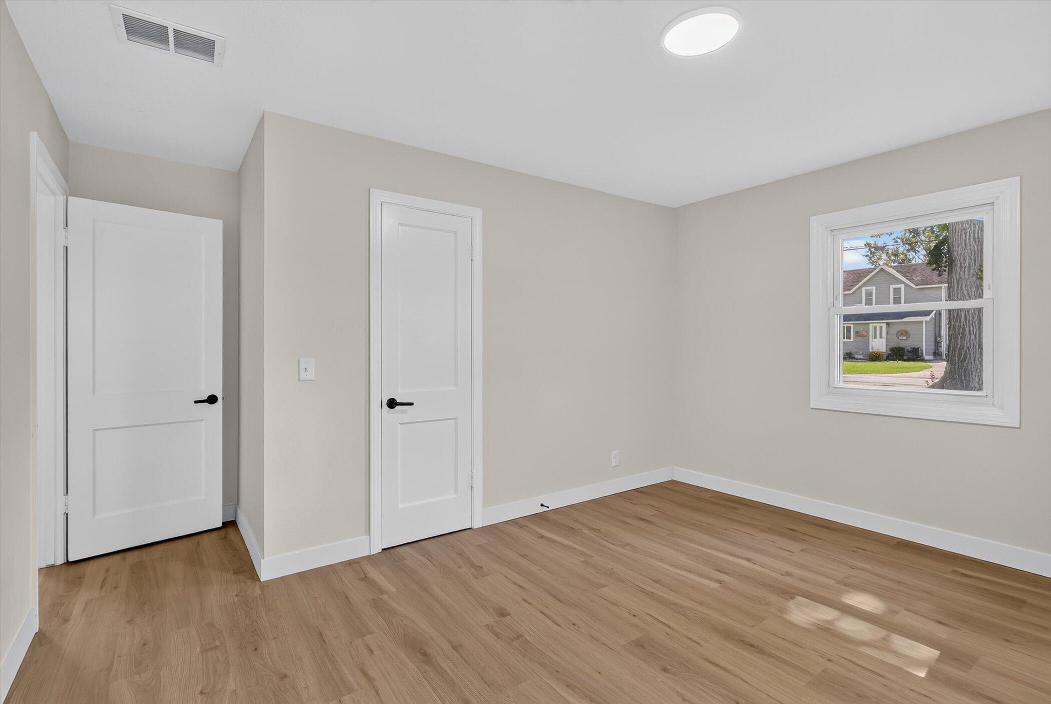 Empty room with light wood floors, two white doors, and a window facing a neighboring house.