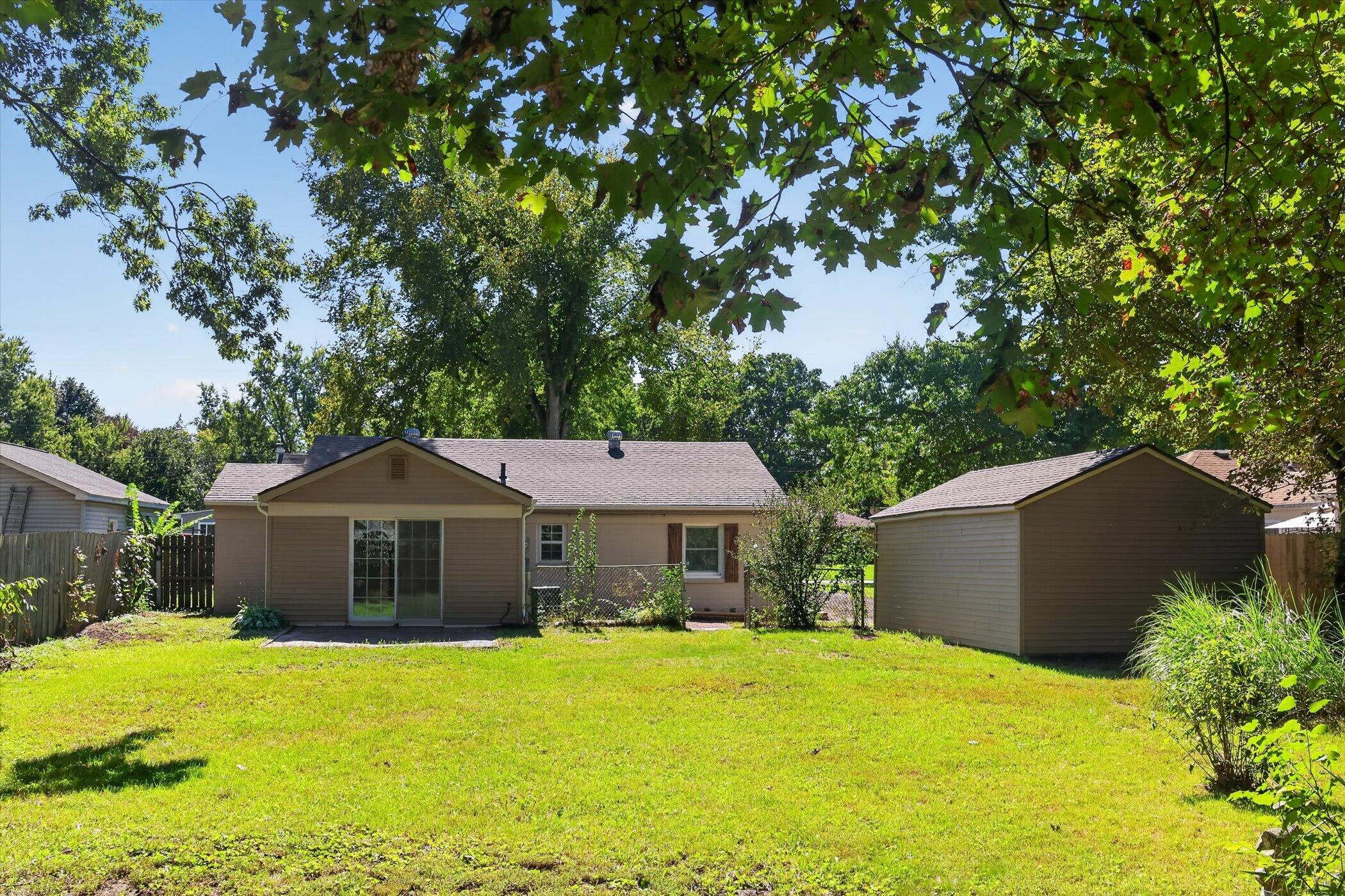 Rear of a tan home with a detached shed and leafy trees surrounding the grassy backyard.