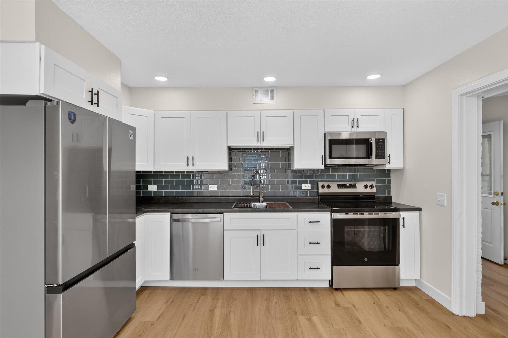 Kitchen with white cabinets, dark tile backsplash, stainless steel appliances, and light wood flooring.