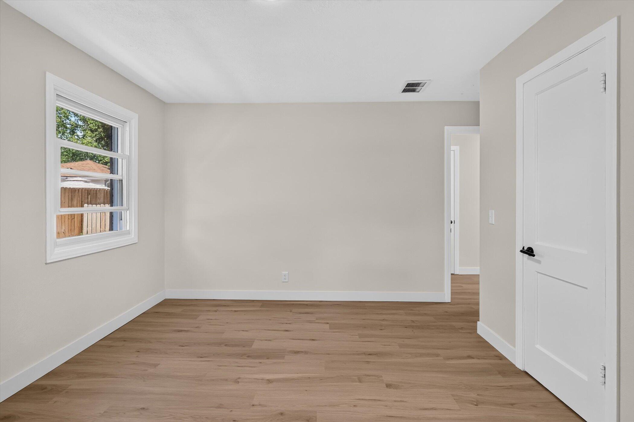 Empty room with warm wood flooring, beige walls, a window, and a white door.