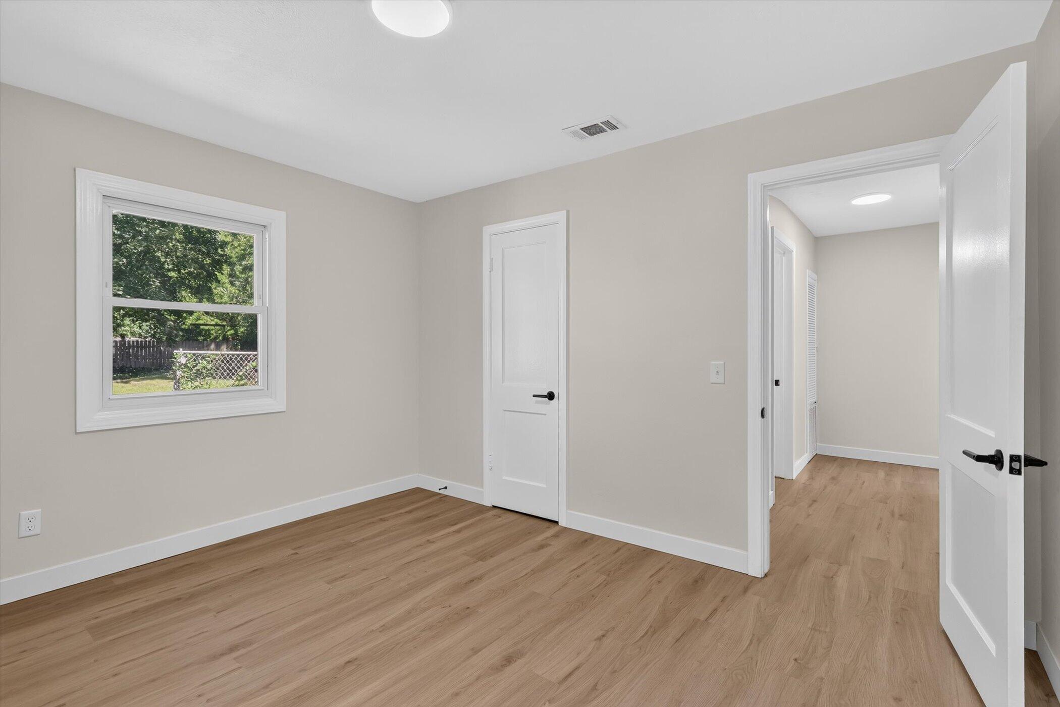 Empty room with light wood floors, a window overlooking a fenced yard, and an open doorway to the hall.