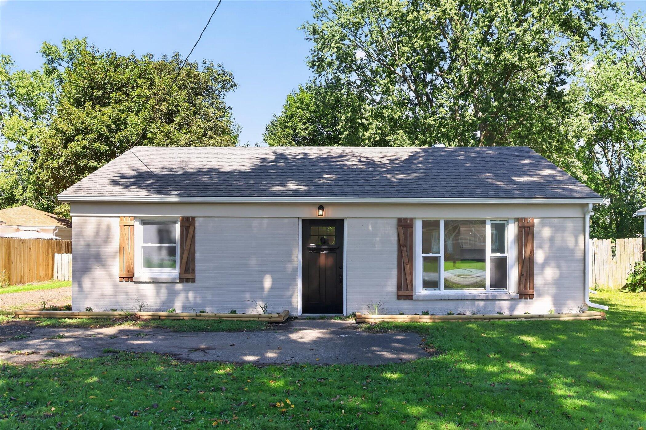 Single-story gray-sided home with wood shutters, a dark front door, and mature trees.